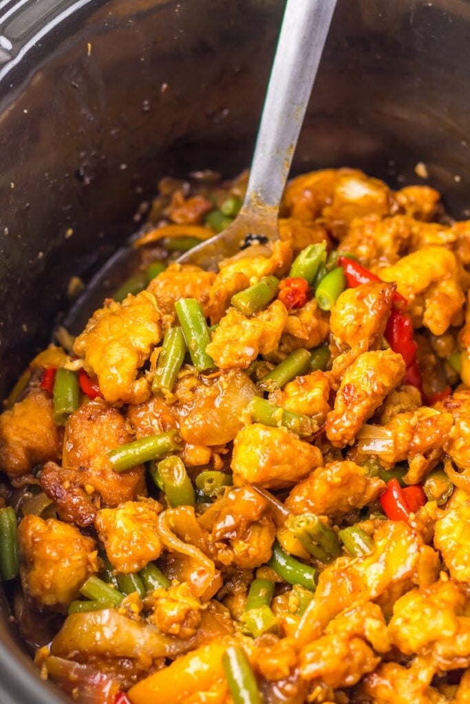 A slow cooker dish with battered chicken pieces, green beans, and red peppers in a glossy sauce, stirred with a metal spoon.