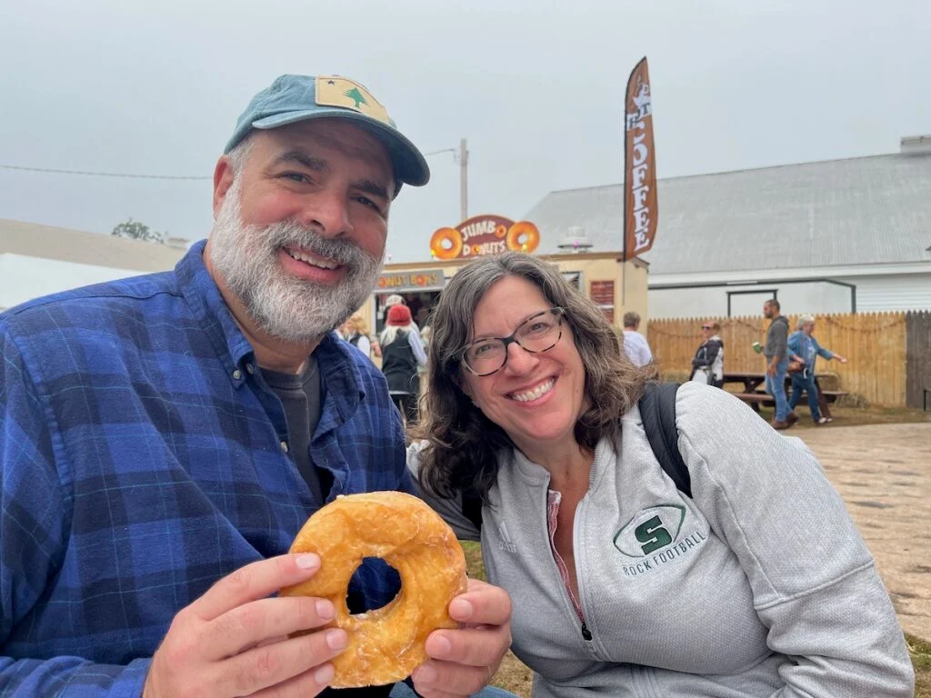 Two people smiling outdoors; the man holds a large donut. There's a sign for coffee and donuts in the background.
