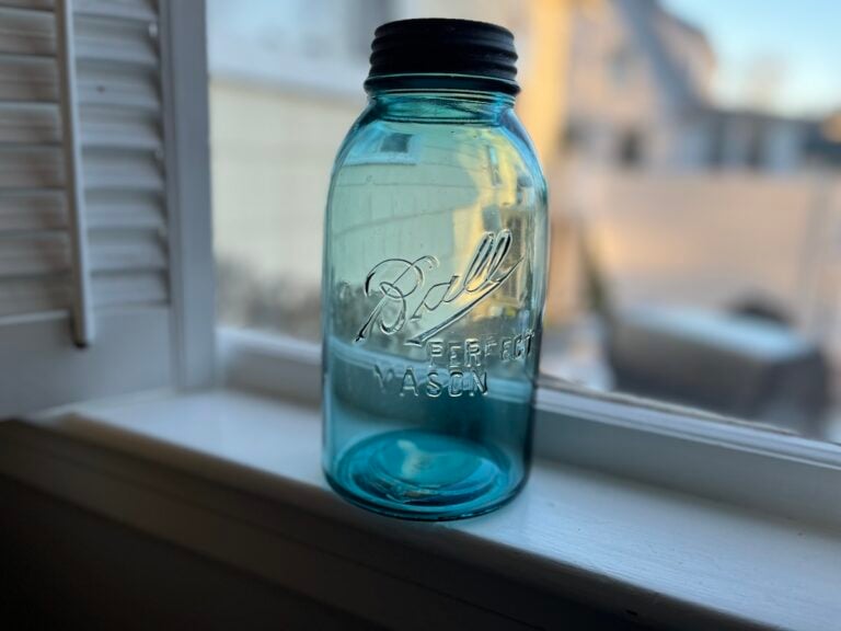 A blue Ball Mason jar with a black lid sits on a windowsill, with sunlight coming through a nearby window.