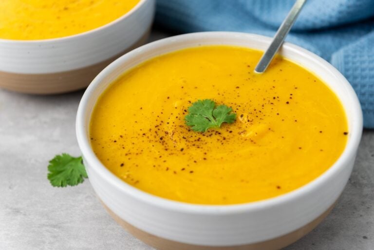 A bowl of bright orange soup garnished with black pepper and a cilantro leaf, accompanied by a spoon.