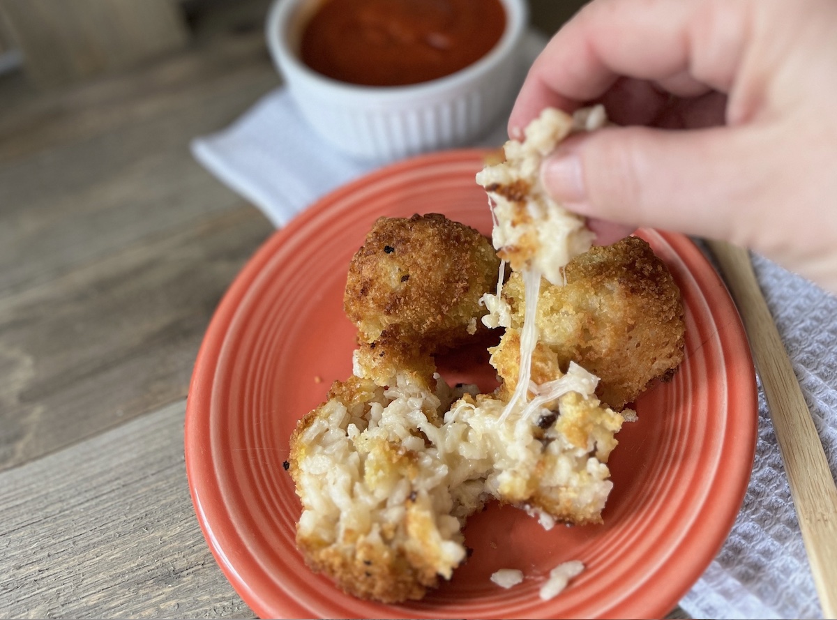 Hand pulling apart a cheesy arancini ball on an orange plate, with a bowl of marinara sauce in the background.