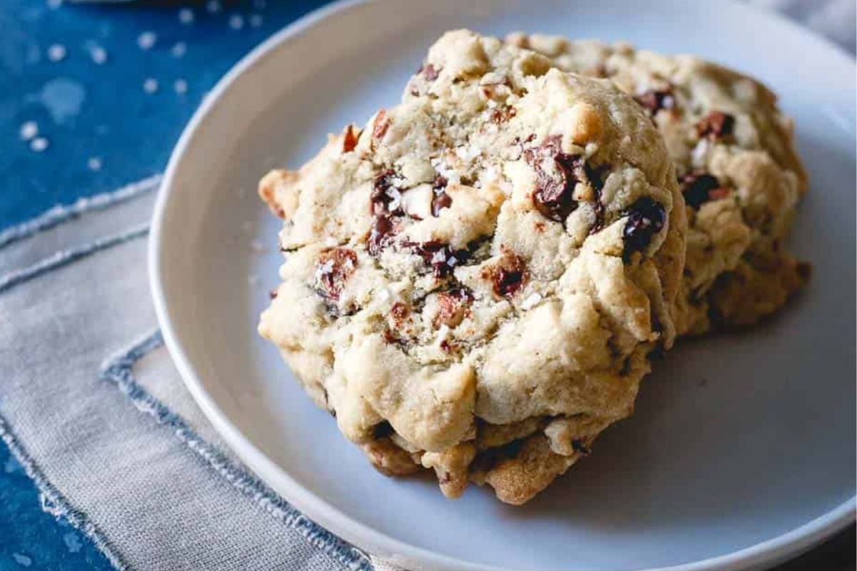 Two chocolate chip cookies on a white plate, placed on a cloth napkin against a blue background.