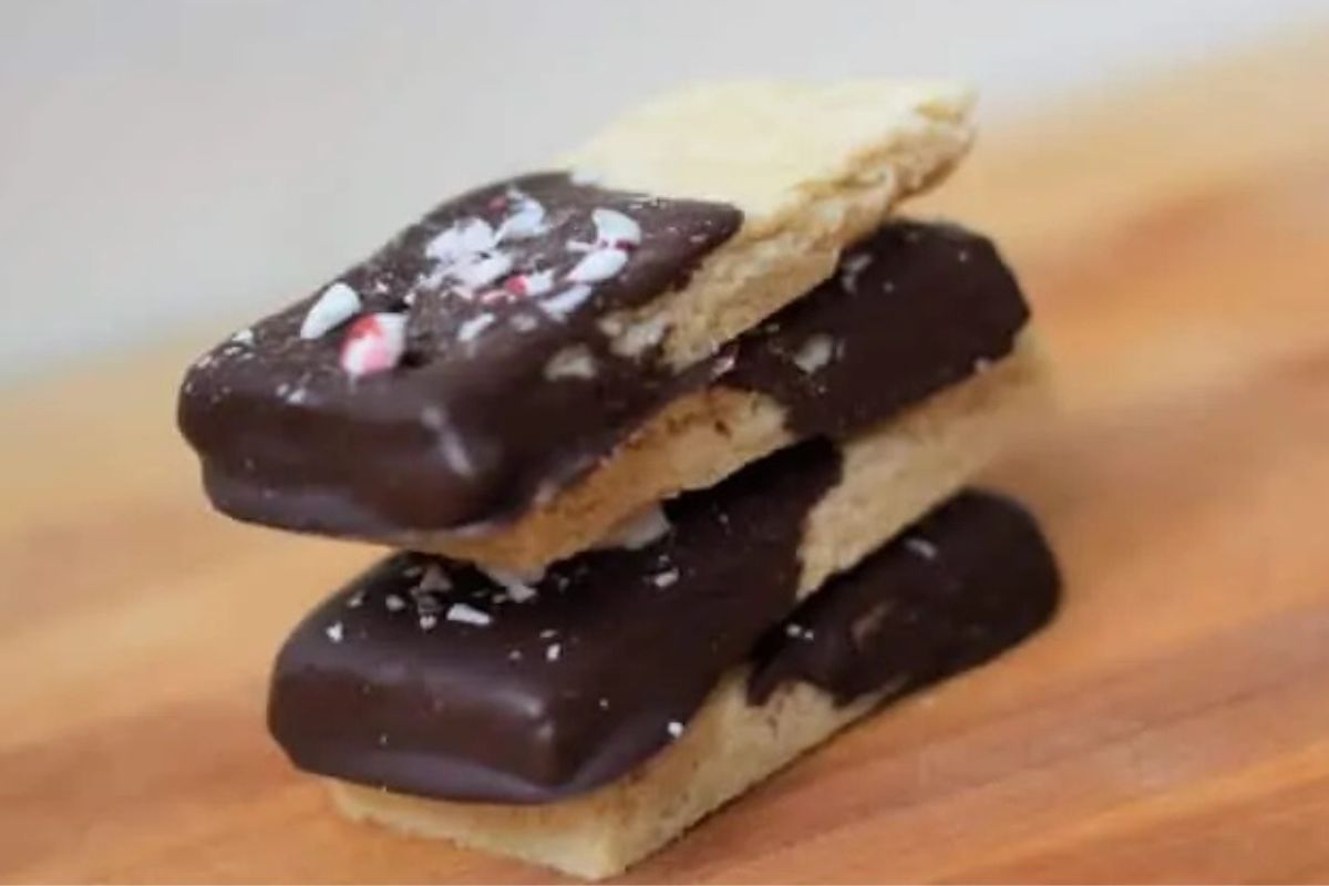 Three shortbread cookies partially dipped in chocolate, stacked on a wooden surface, with peppermint pieces on top.