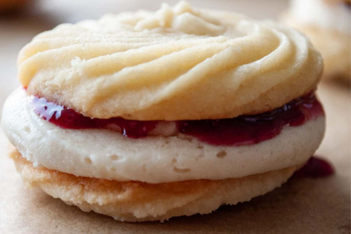 Close-up of a sandwich cookie with cream and raspberry jam filling between two buttery shortbread layers.