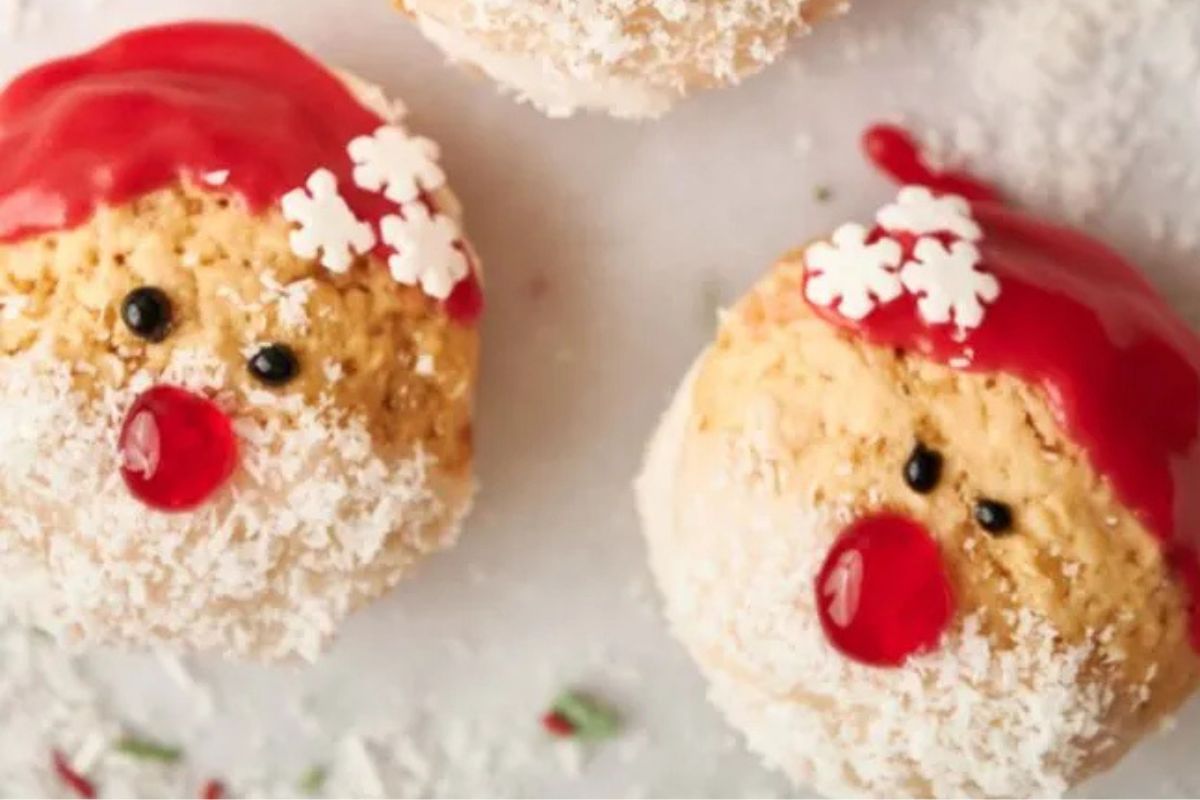 Two Christmas-themed cupcakes decorated with red icing hats, white snowflakes, coconut flakes for beards, and red and black icing for eyes and noses on a light surface.