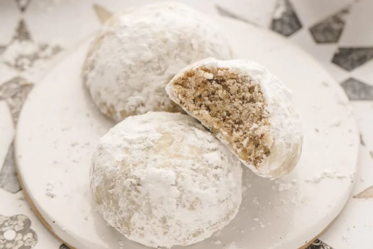 Two powdered sugar cookies on a plate, one partially bitten to reveal a crumbly interior.