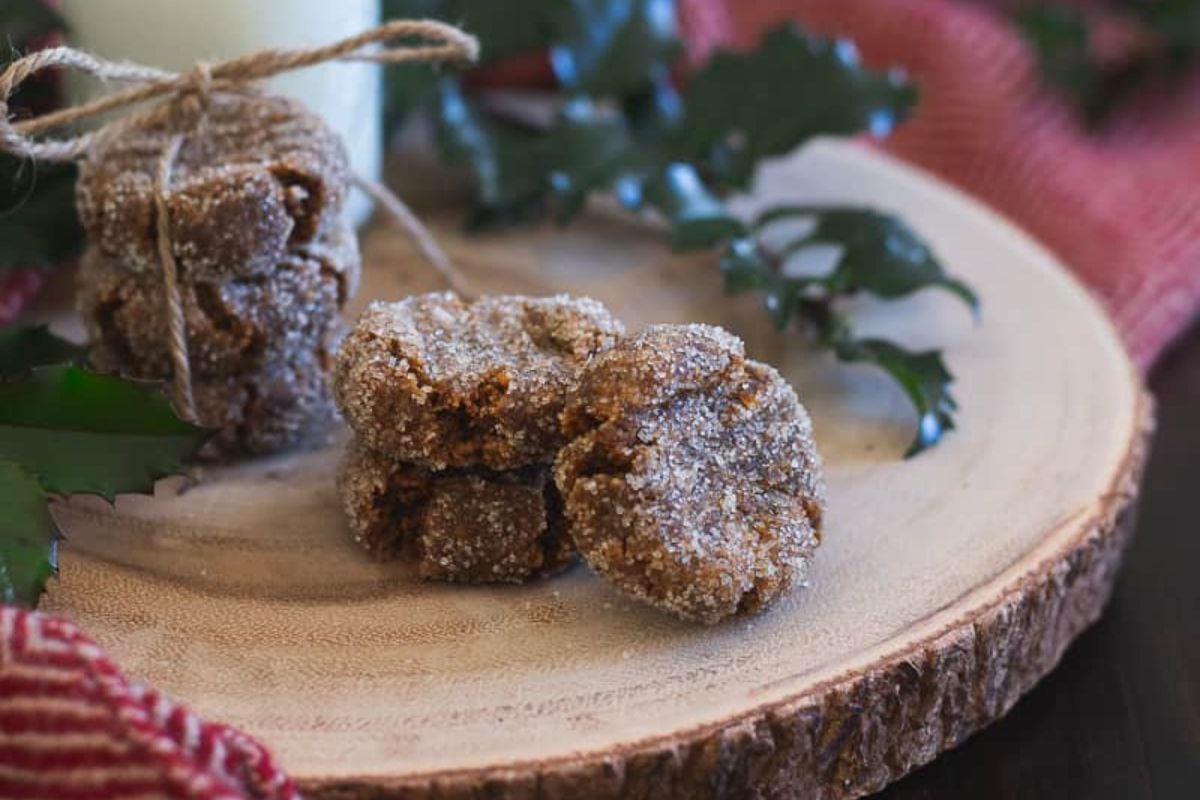 Sugared cookies are stacked on a wooden platter, surrounded by holly leaves and a red cloth.