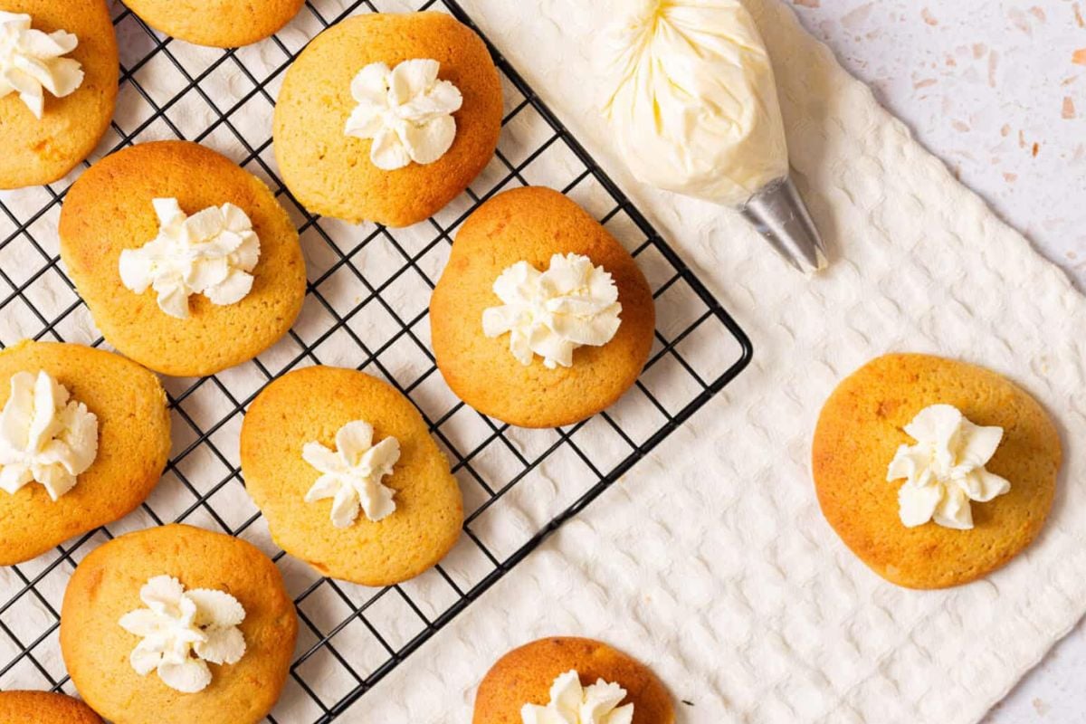 Cookies with white frosting on top sit on a cooling rack and a white cloth, with a piping bag nearby.