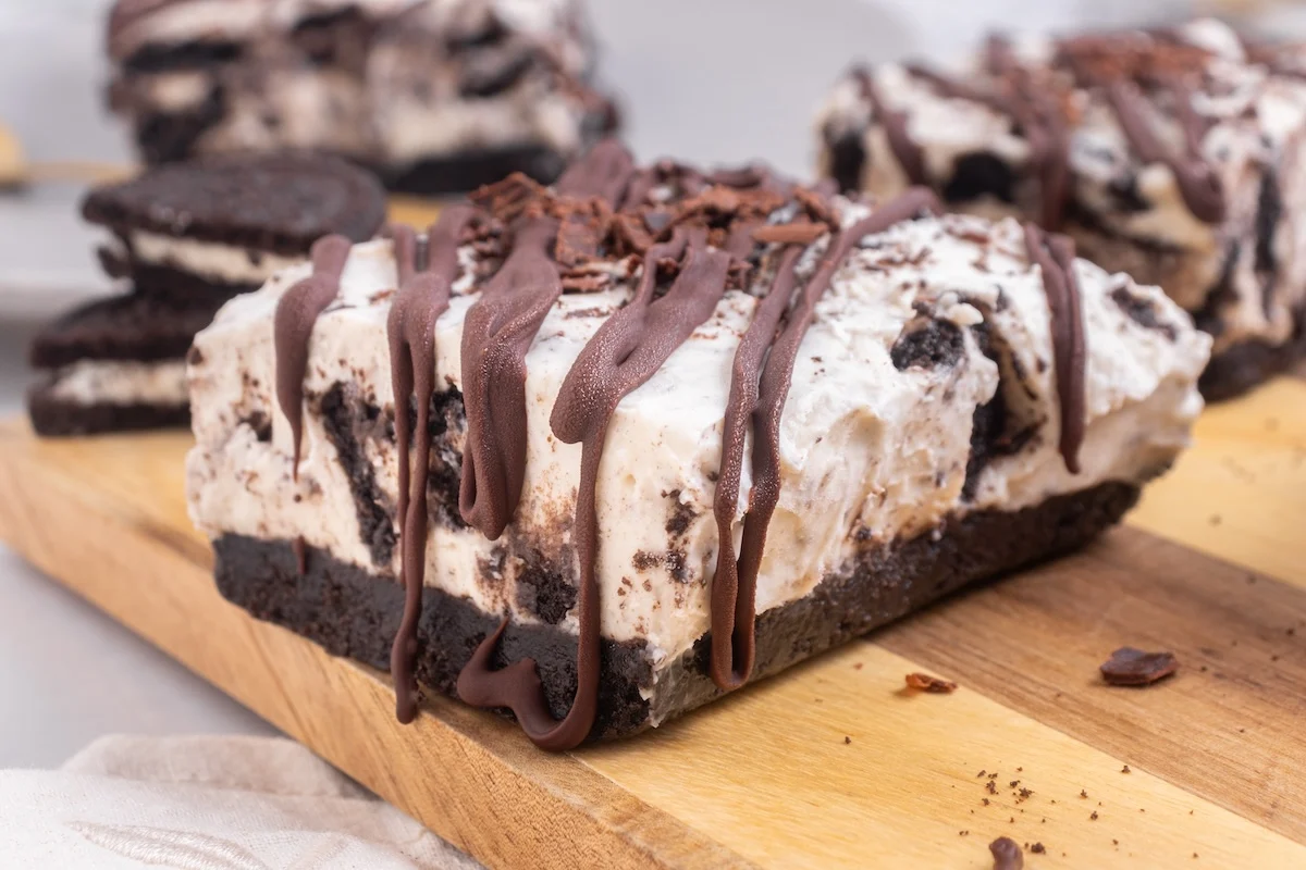 Close-up of a dessert bar with layers of cream and chocolate cookie pieces, topped with chocolate drizzle.