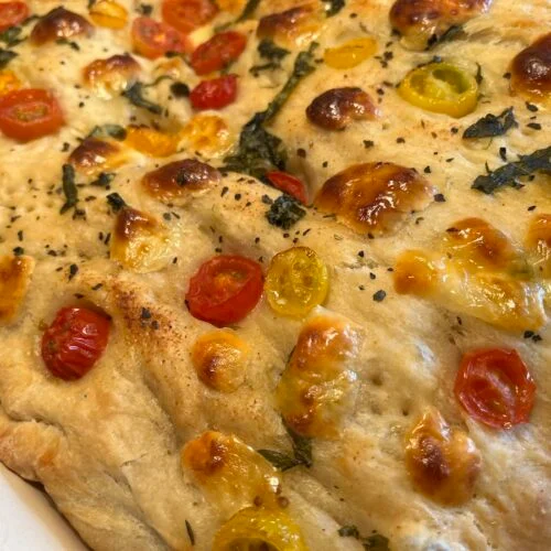 Close-up of baked focaccia topped with cherry tomatoes, melted cheese, and herbs.