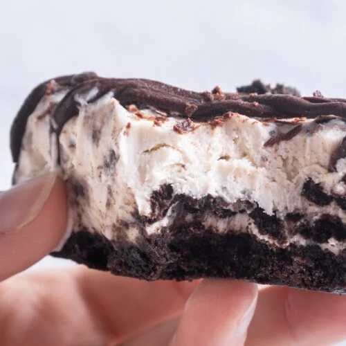 Close-up of a hand holding a partially eaten dessert with layers of chocolate crust, creamy filling, and chocolate drizzle.