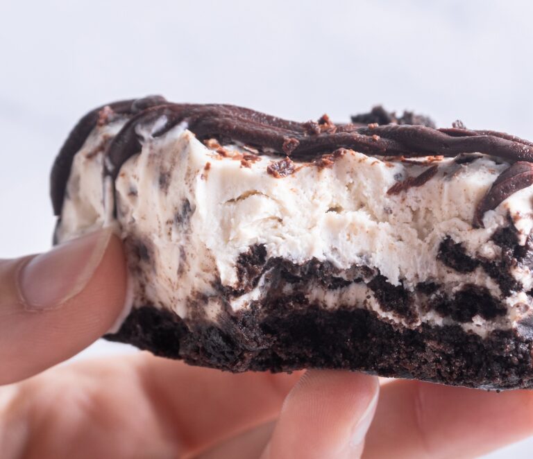 Close-up of a hand holding a partially eaten dessert with layers of chocolate crust, creamy filling, and chocolate drizzle.