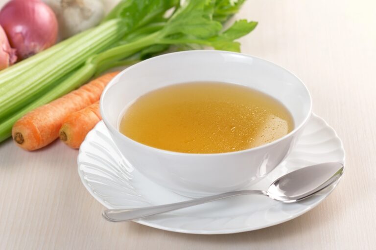 A white bowl of clear DIY vegetable broth sits on a saucer with a spoon beside it. In the background, fresh carrots, celery stalks, and a red onion hint at the homemade goodness within.