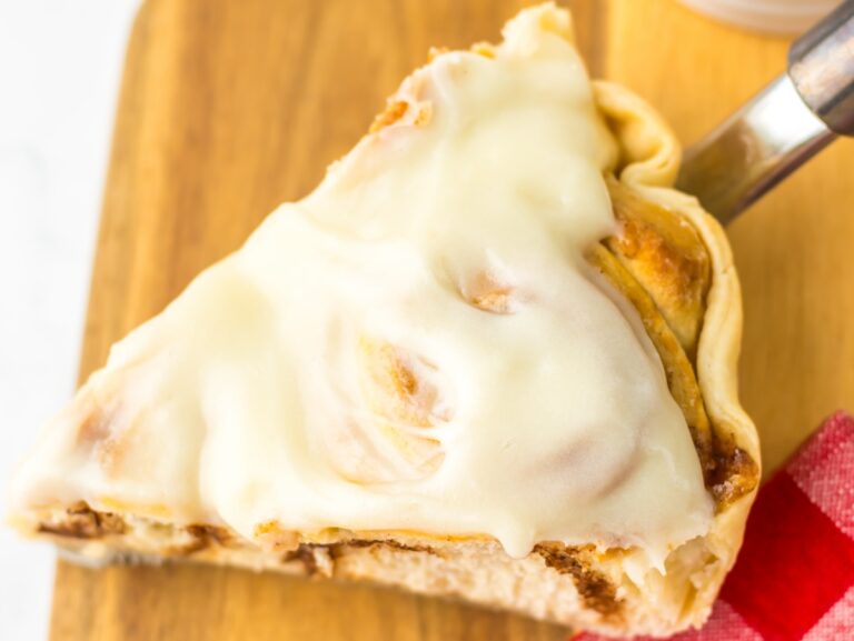 A slice of cinnamon roll pie topped with white icing on a wooden board. A pie server is partially visible under the slice.