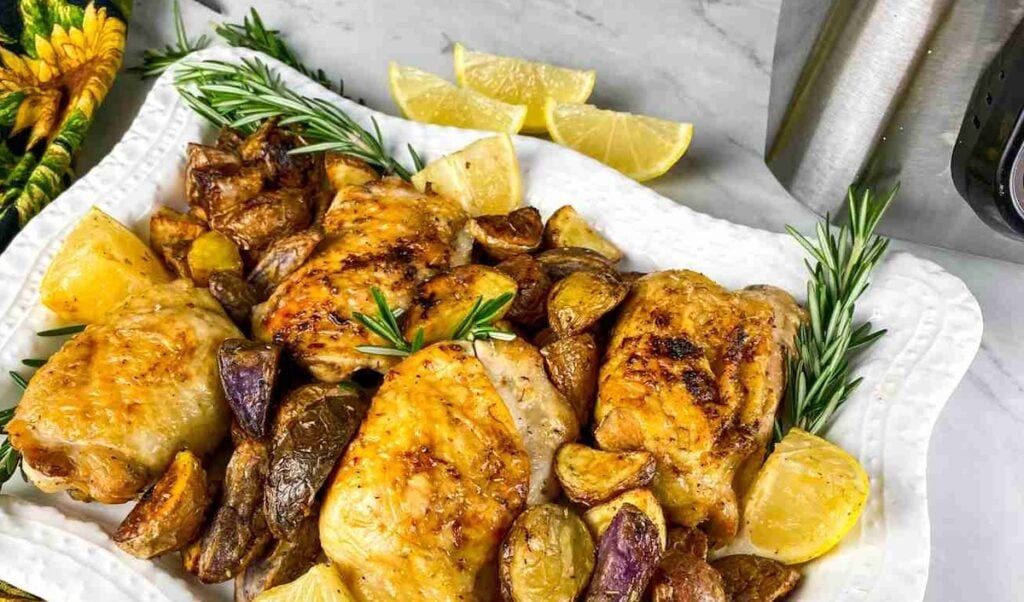 Roasted chicken thighs with potatoes and lemon wedges, garnished with rosemary, on a white platter.