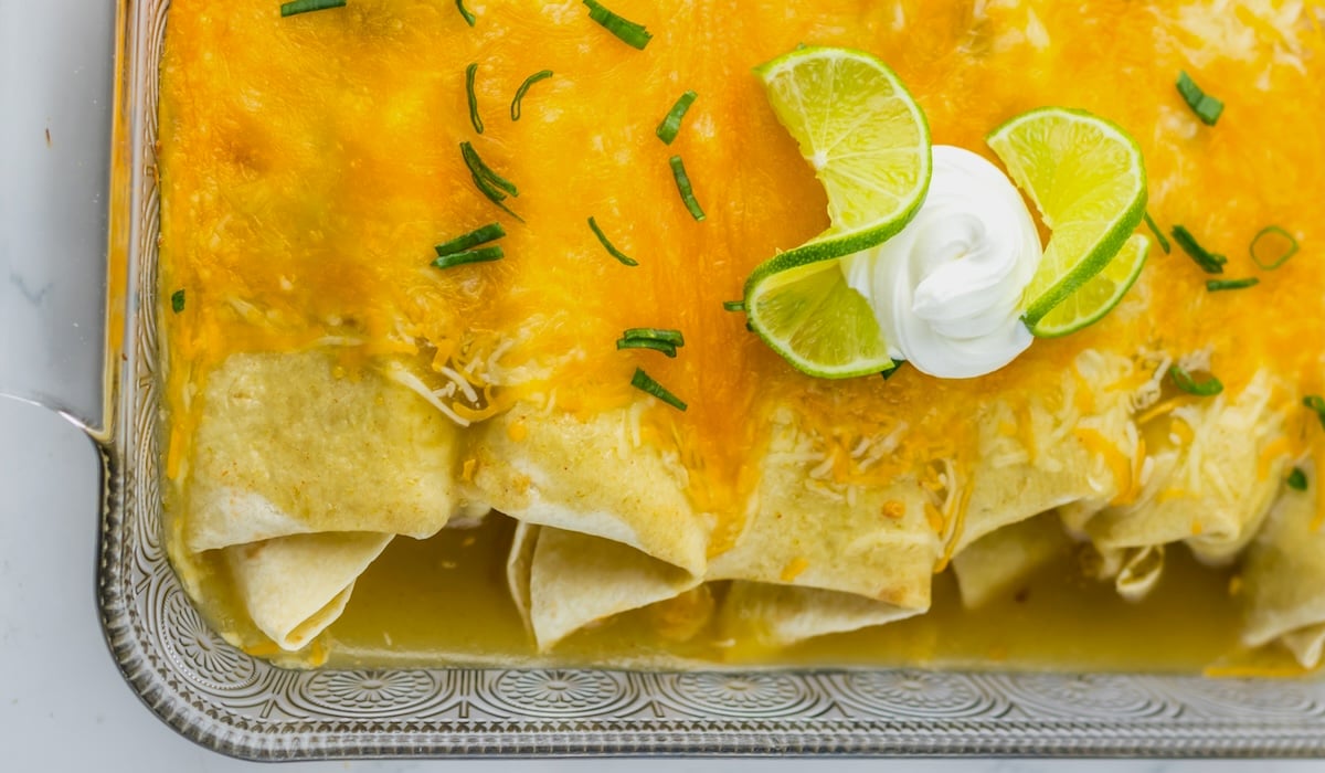 A dish of enchiladas filled with slow cooker honey lime shredded chicken, topped with melted cheese, lime slices, sour cream, and chives.