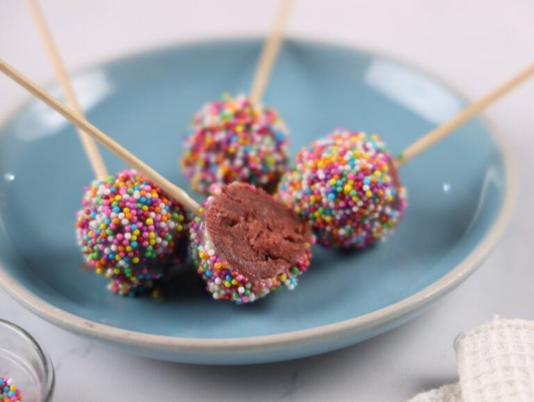 Four cake pops coated with colorful sprinkles are on a blue plate. One pop is bitten in half, revealing its interior. Wooden sticks are inserted into each pop.