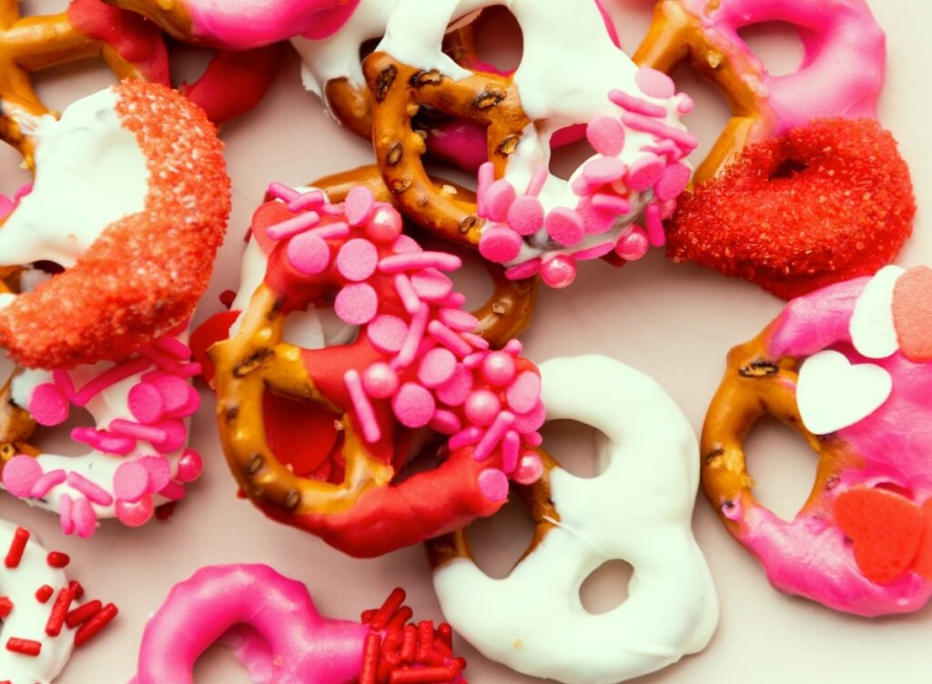 Assorted pretzels covered in red, pink, and white icing with various sprinkles, placed on a light surface.