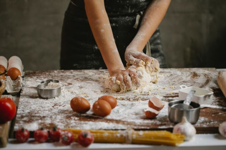 Person kneading dough on a floured wooden surface surrounded by eggs, tomatoes, and pasta.