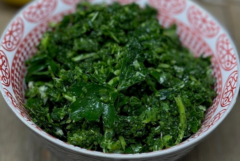 A bowl of finely chopped green herbs sits in a red and white decorative bowl on a wooden surface.