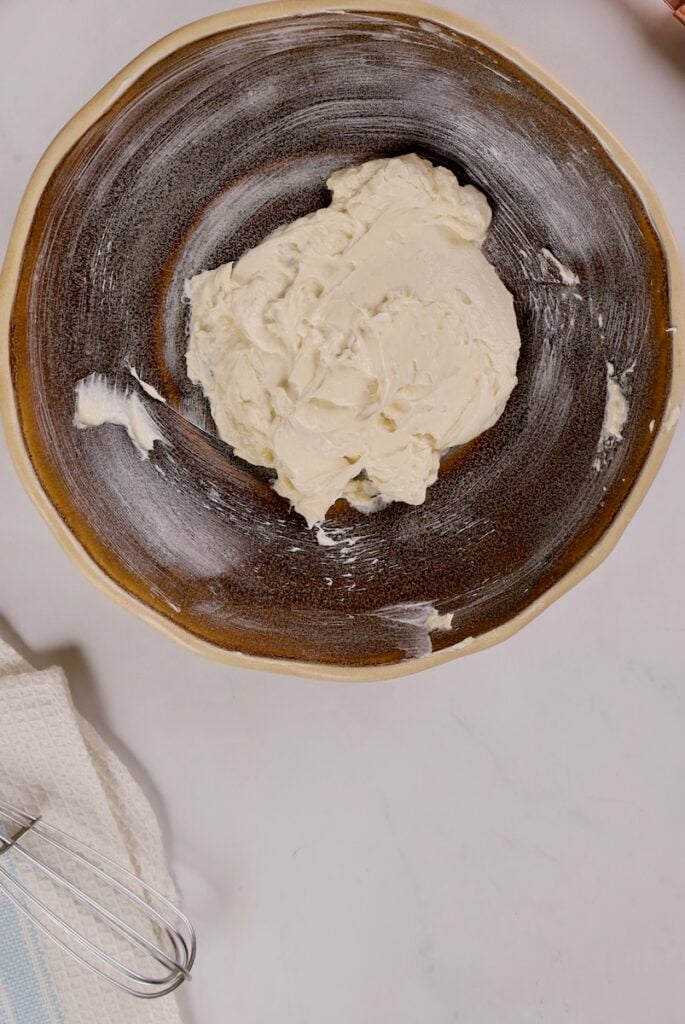 A ceramic bowl with cream cheese on a white surface sits beside a whisk and towel, hinting at the sweet delights like cake pops in a mug waiting to be created.