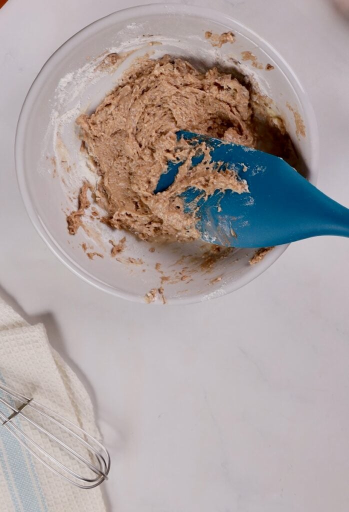 A bowl with dough mixture and a blue spatula on a white surface hints at cake pops in a mug in the making. Nearby, a whisk and cloth stand ready for their part.