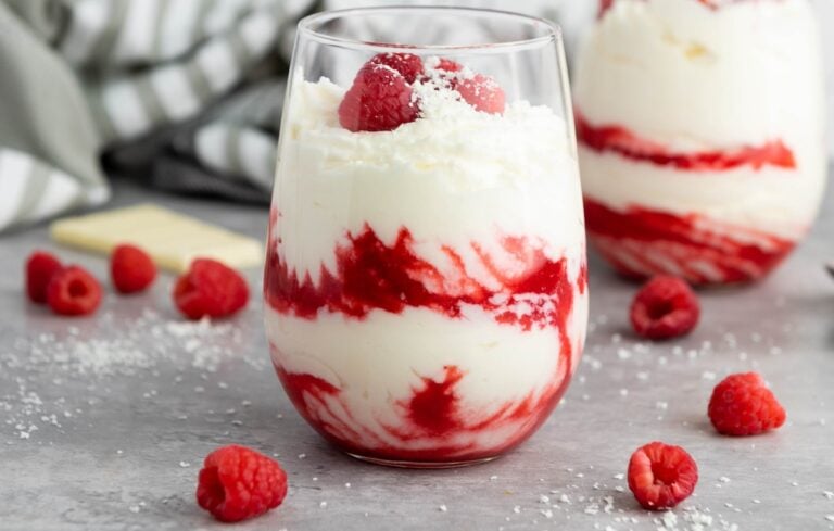 Glass of parfait with creamy layers, raspberry swirls, and topped with raspberries and white chocolate flakes, surrounded by scattered raspberries on a gray surface.