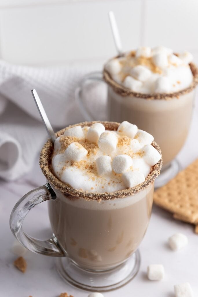 Two clear glass mugs of hot chocolate topped with whipped cream, mini marshmallows, and crushed graham crackers; silver spoons inside. White towel and graham crackers in the background.