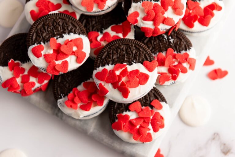 White chocolate-covered Oreos partially dipped in white icing, topped with red heart sprinkles, are beautifully arranged on a pristine white surface.