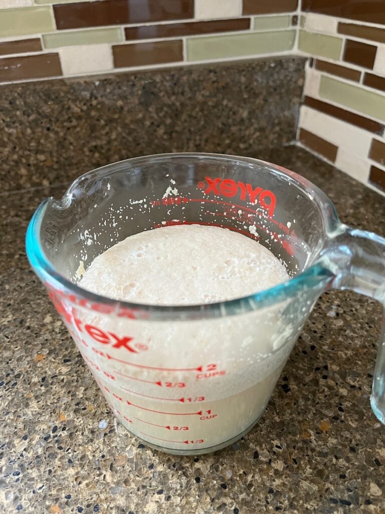 A clear Pyrex measuring cup filled with a frothy, light beige liquid sits on a speckled countertop, ready to be transformed into rustic crock pot sourdough bread, with a tiled backsplash in the background.