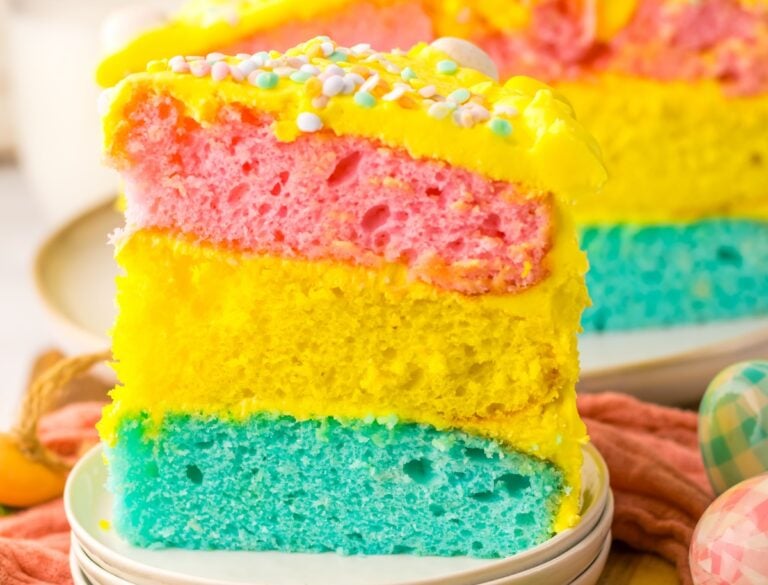 A slice of colorful layer cake with pink, yellow, and blue layers, topped with yellow frosting and sprinkles.