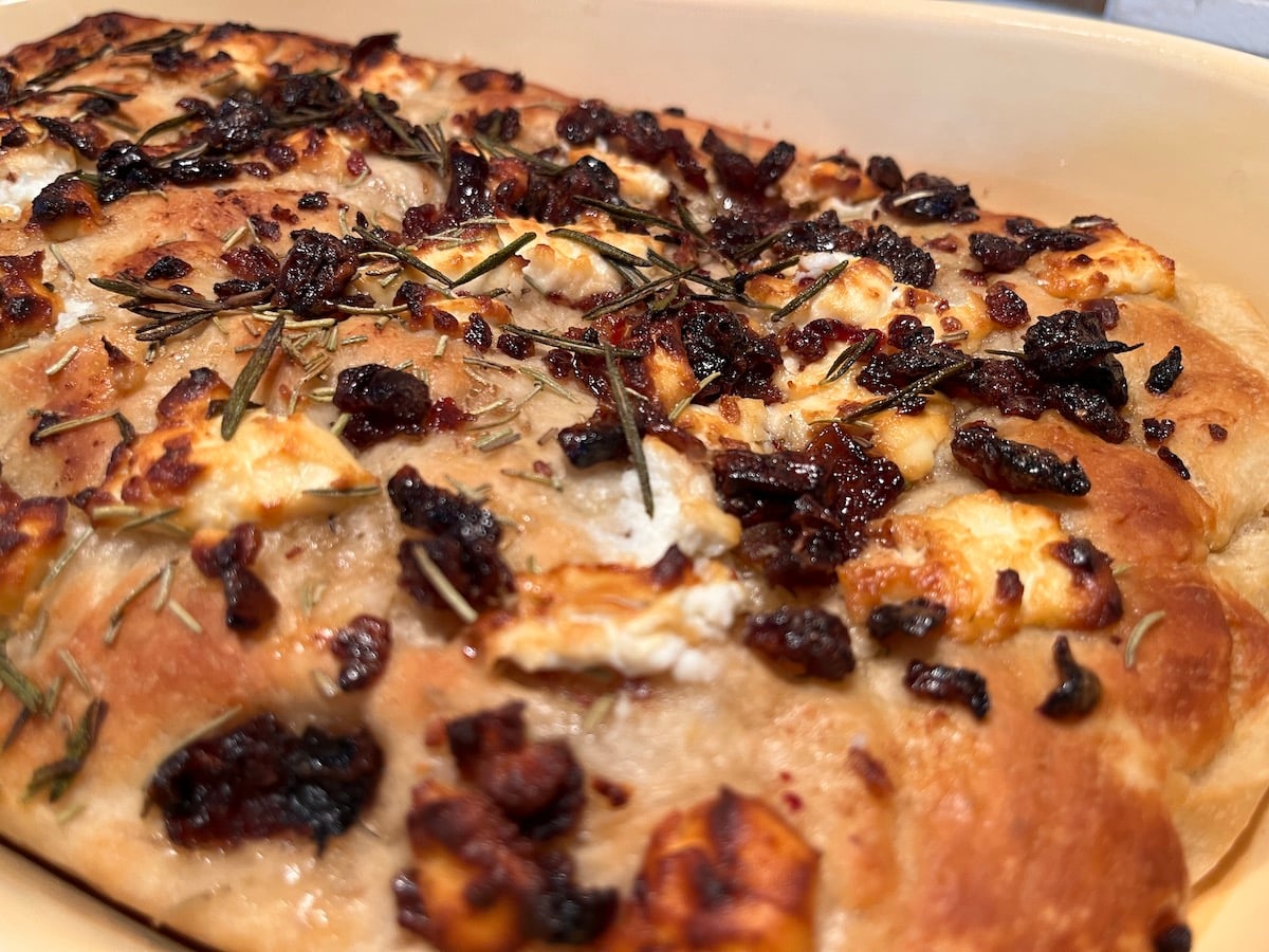 Close-up of a homemade focaccia topped with caramelized onions, rosemary, and chunks of cheese in a baking dish.