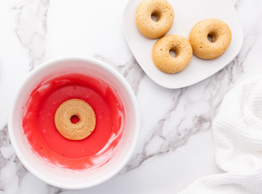 A doughnut being dipped into red icing inside a white bowl, with three undipped doughnuts on a white plate next to it.