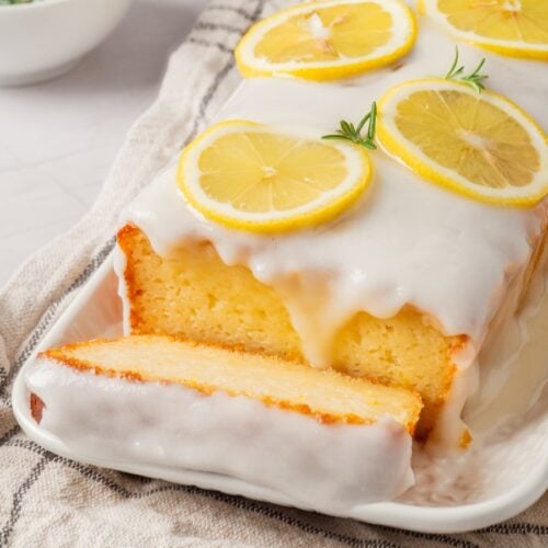 Lemon loaf cake with white icing and lemon slices on top, served on a rectangular plate with a piece cut.