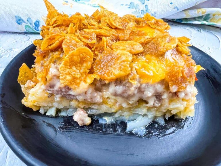 A slice of casserole on a black plate, topped with crispy corn flakes, showcases layers of ground meat, cheese, and potatoes—perfect for those who love breakfast for dinner recipes.