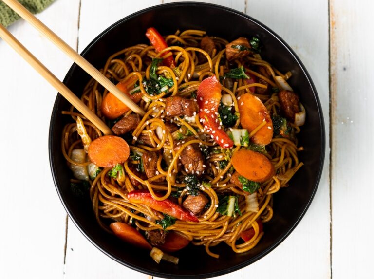 A bowl of stir-fried noodles with vegetables, including carrots and bell peppers, topped with sesame seeds, rests alongside an instant pot pork lo mein. Chopsticks are elegantly perched on the bowl.