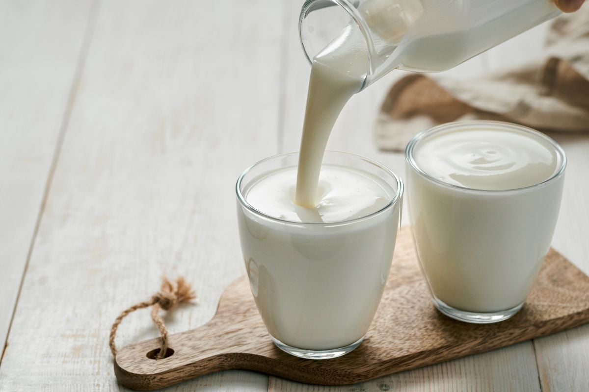 Milk being poured from a glass jug into a glass on a wooden board, next to another glass of milk and some egg alternatives, on a light wooden surface.