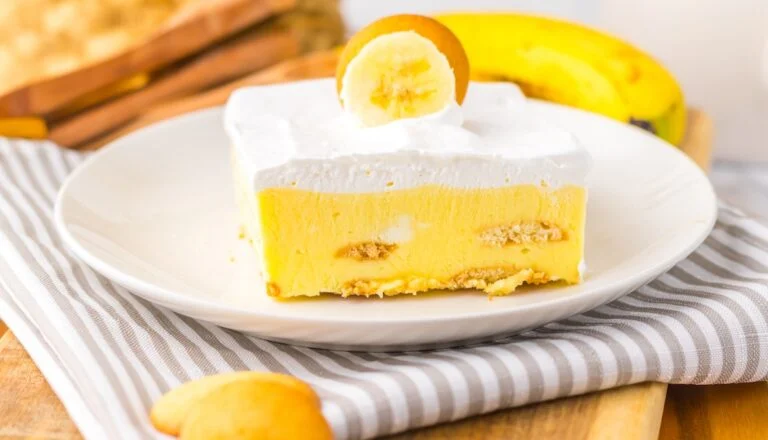 A slice of banana pudding is topped with whipped cream and a banana slice, served on a white plate.