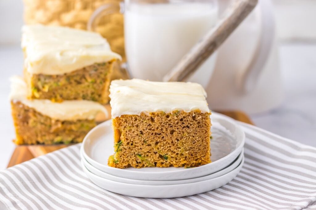 Slice of frosted zucchini cake on stacked plates with additional cake slices and a glass of milk in the background.