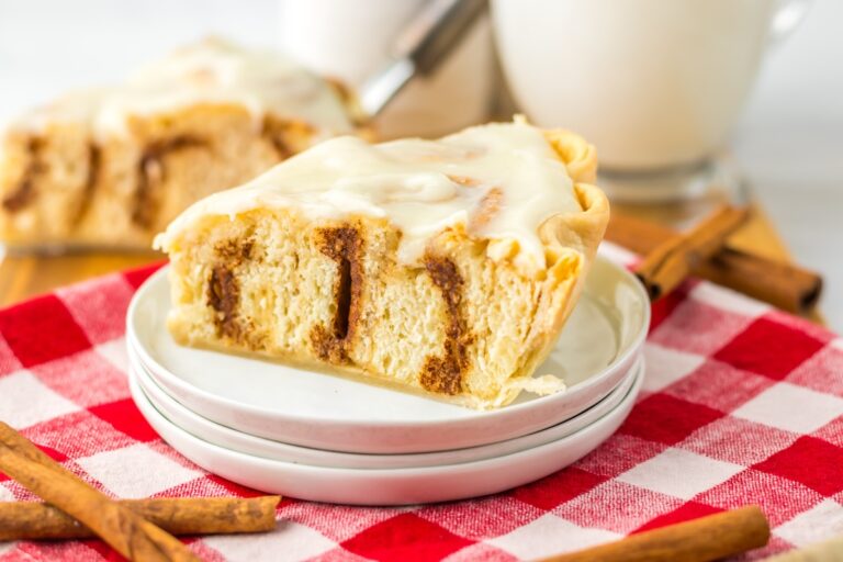 A slice of cinnamon roll cheesecake on a white plate. The cheesecake is topped with icing and sits on a red and white checkered cloth. Cinnamon sticks are nearby.