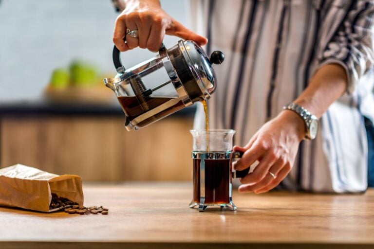 A person skillfully demonstrates how to make French press coffee, pouring the rich brew into a glass on a wooden counter, with a bag of coffee beans close at hand.