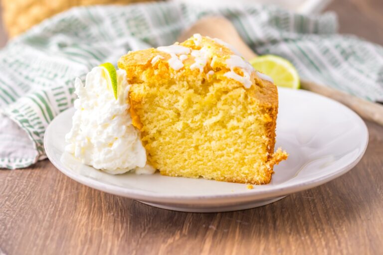 Slice of lemon cake with white icing on a plate, accompanied by a dollop of whipped cream and a lime slice.