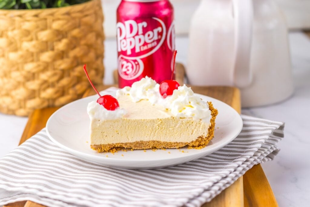 A slice of cream pie topped with whipped cream and cherries on a white plate. A can of Dr Pepper and a basket are in the background.