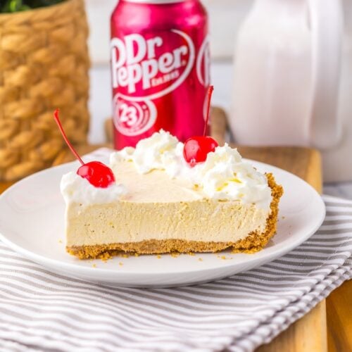 A slice of cream pie topped with whipped cream and cherries on a white plate. A can of Dr Pepper and a basket are in the background.