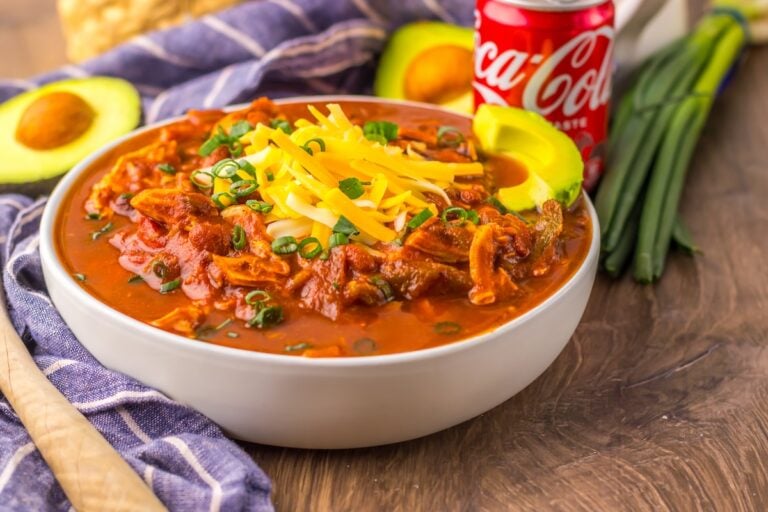 A bowl of chili topped with shredded cheese and green onions, surrounded by avocado slices and a can of Coca-Cola, on a wooden table with a blue cloth.