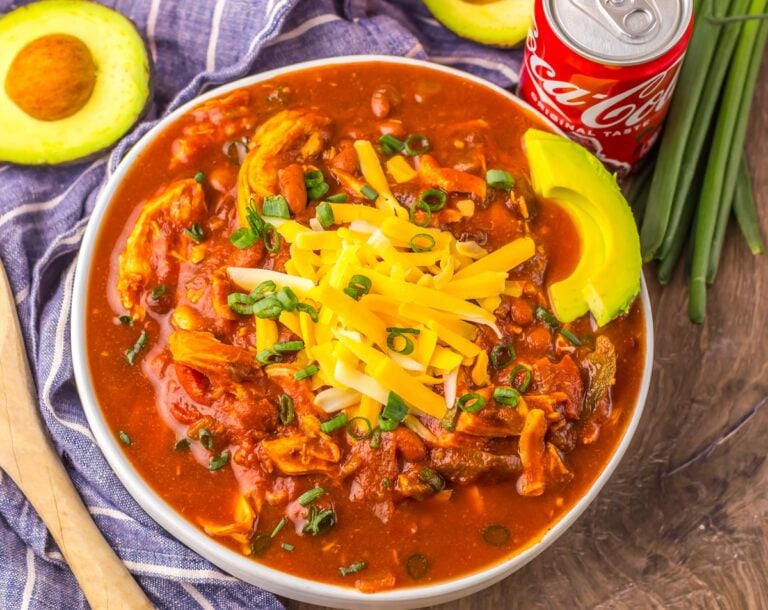A bowl of chili topped with shredded cheese and green onions, garnished with avocado slices. A can of Coca-Cola and halved avocado are next to the bowl.