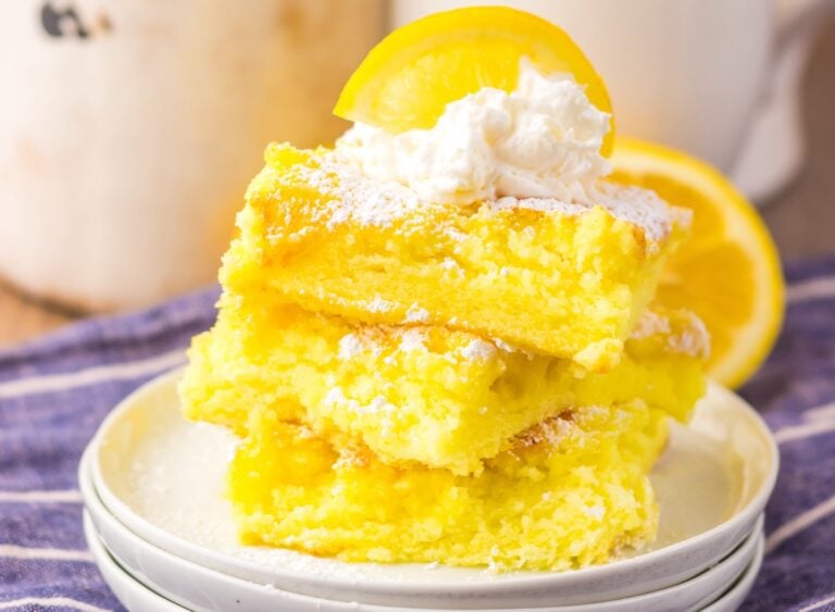 A stack of lemon bars topped with whipped cream and a lemon slice on a white plate, placed on a blue striped cloth.