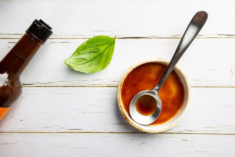 A spoon rests in a small bowl of brown sauce on a white wooden surface, accompanied by a green leaf and the top portion of a brown sauce bottle.