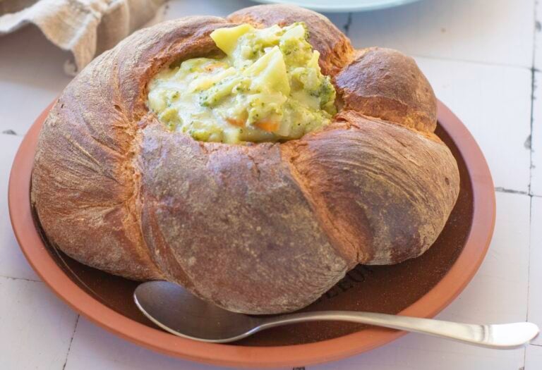 A round bread bowl filled with creamy soup containing chunks of broccoli and potatoes, placed on a brown plate with a spoon beside it.