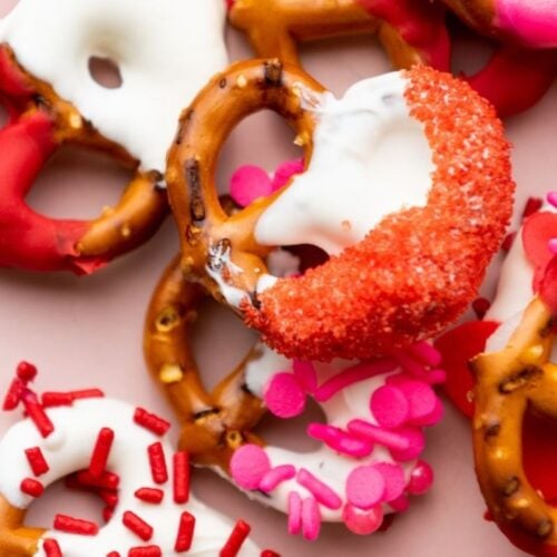 A pile of pretzels coated with white, red, and pink chocolate and decorated with sprinkles on a light pink background.